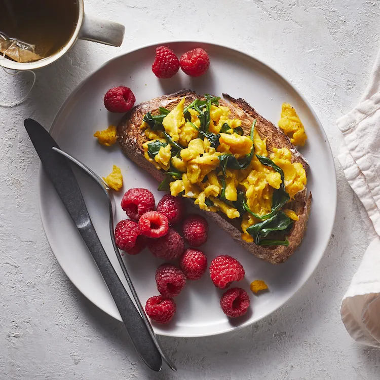 A healthy meal consisting of buttered bread with scrambled eggs, spinach with raspberries and green tea